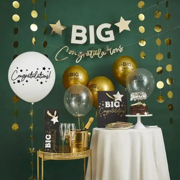 Green and gold celebration display with balloons, hanging decorations, champagne, cake, and Big Congratulations signage on a party table.